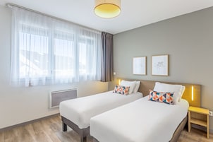 Habitación de hotel con dos camas, una ventana con cortinas translúcidas, un radiador y una mesita de noche con lámpara.