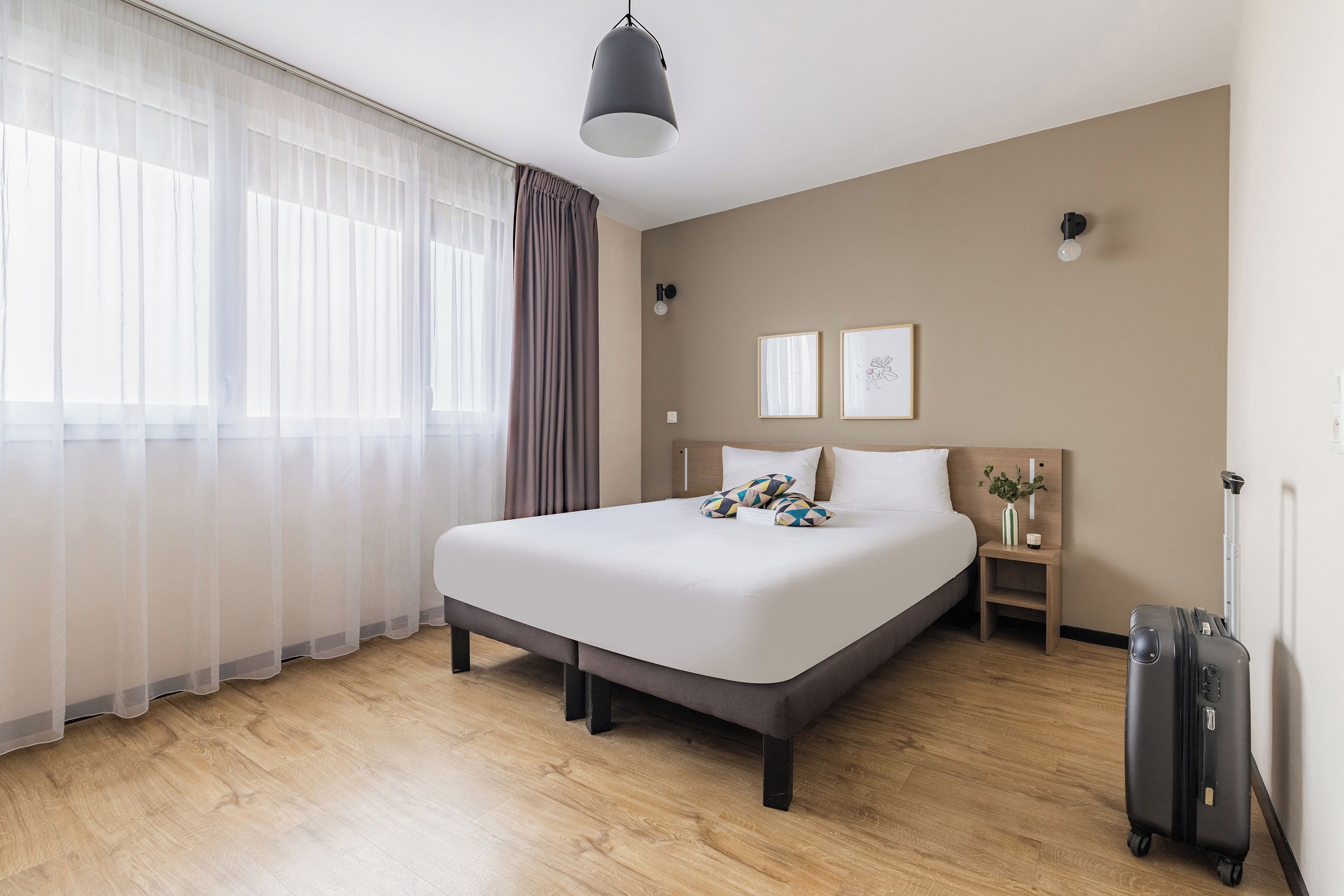Quarto de hotel com uma cama grande, mesa de cabeceira com abajur, mala e janela com cortinas leves.