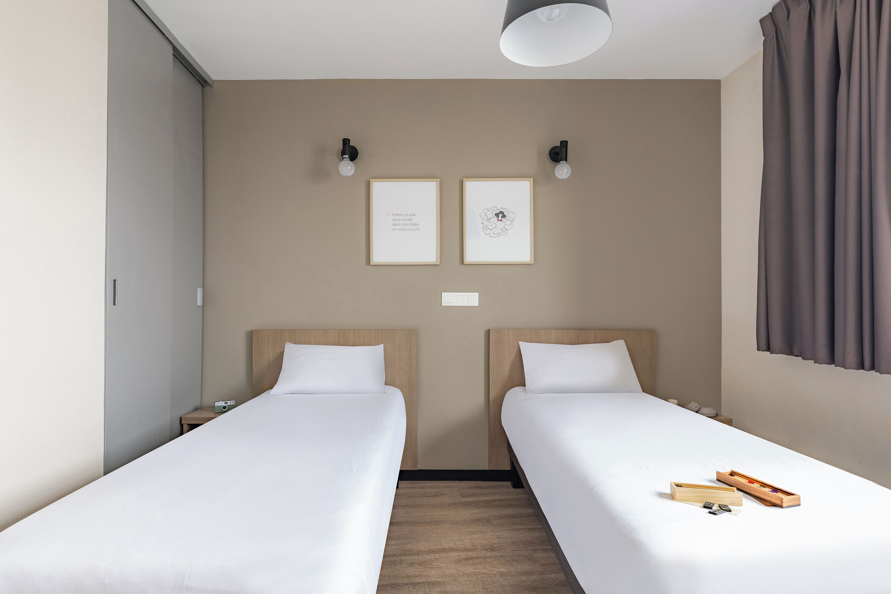 Une chambre d’hôtel avec deux lits simples, une tête de lit en bois, une petite table de chevet avec une lampe et un luminaire fixé au mur.