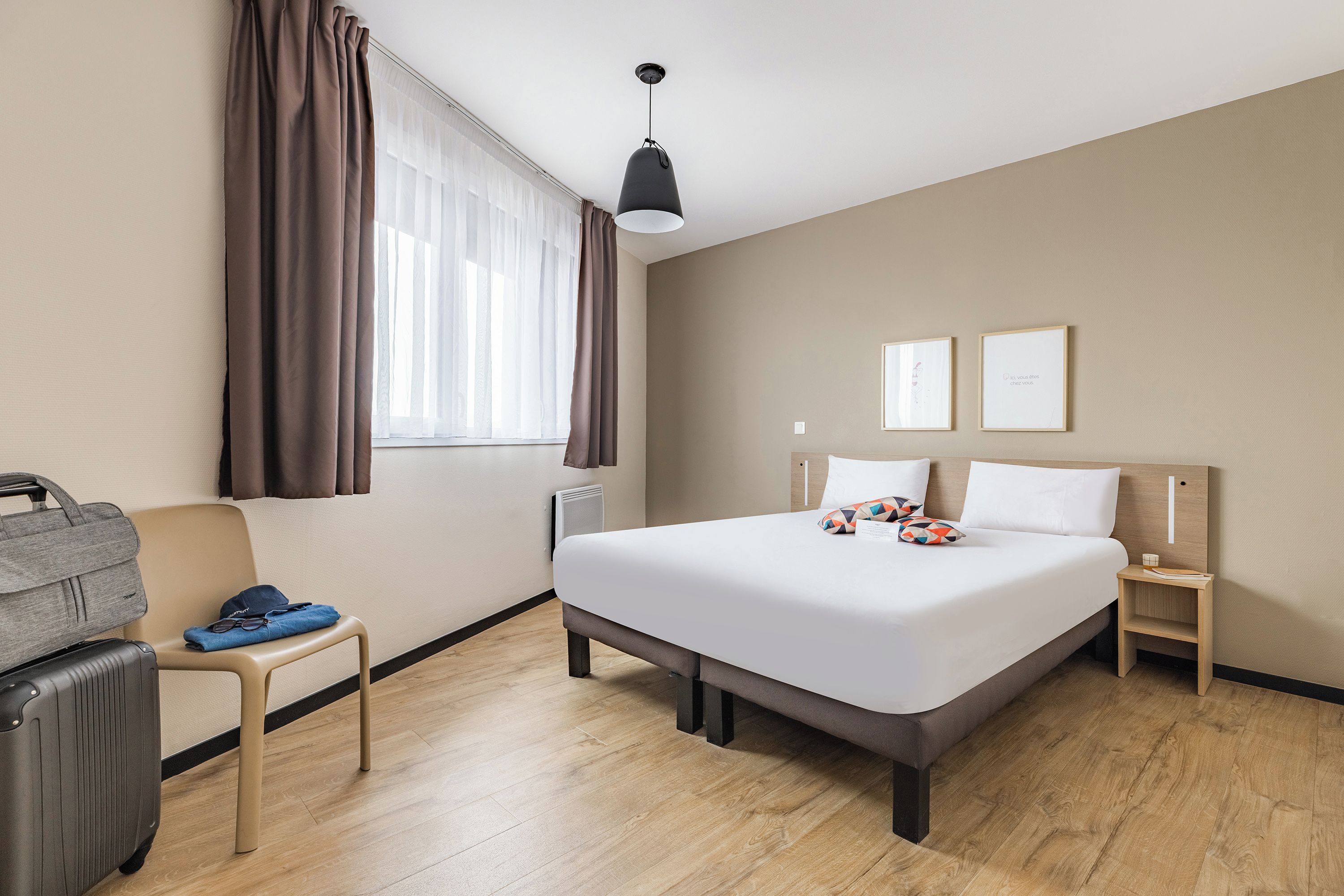 A hotel room with a bed, a chair, a suitcase, a nightstand, and a window with curtains.