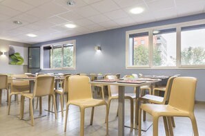 Un área de comedor moderna con sillas y mesas color beige, ventanas amplias y piso de baldosas.