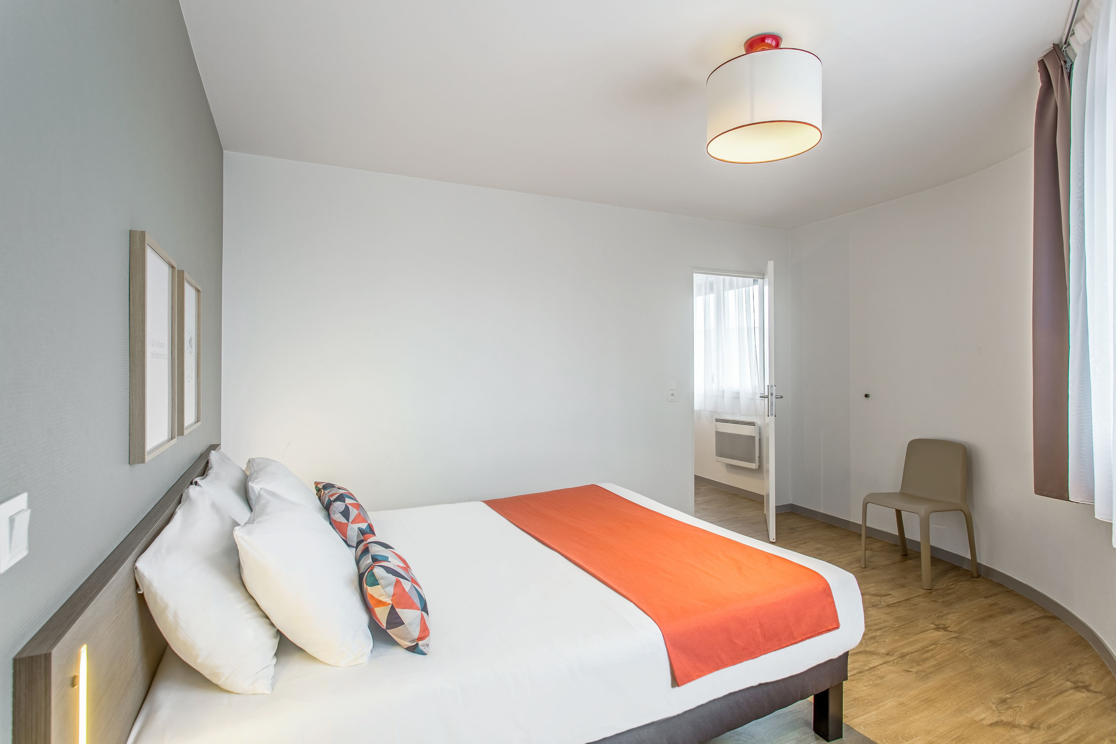 A modern hotel room with a large bed, a bedside table, a chair, and a window with curtains.