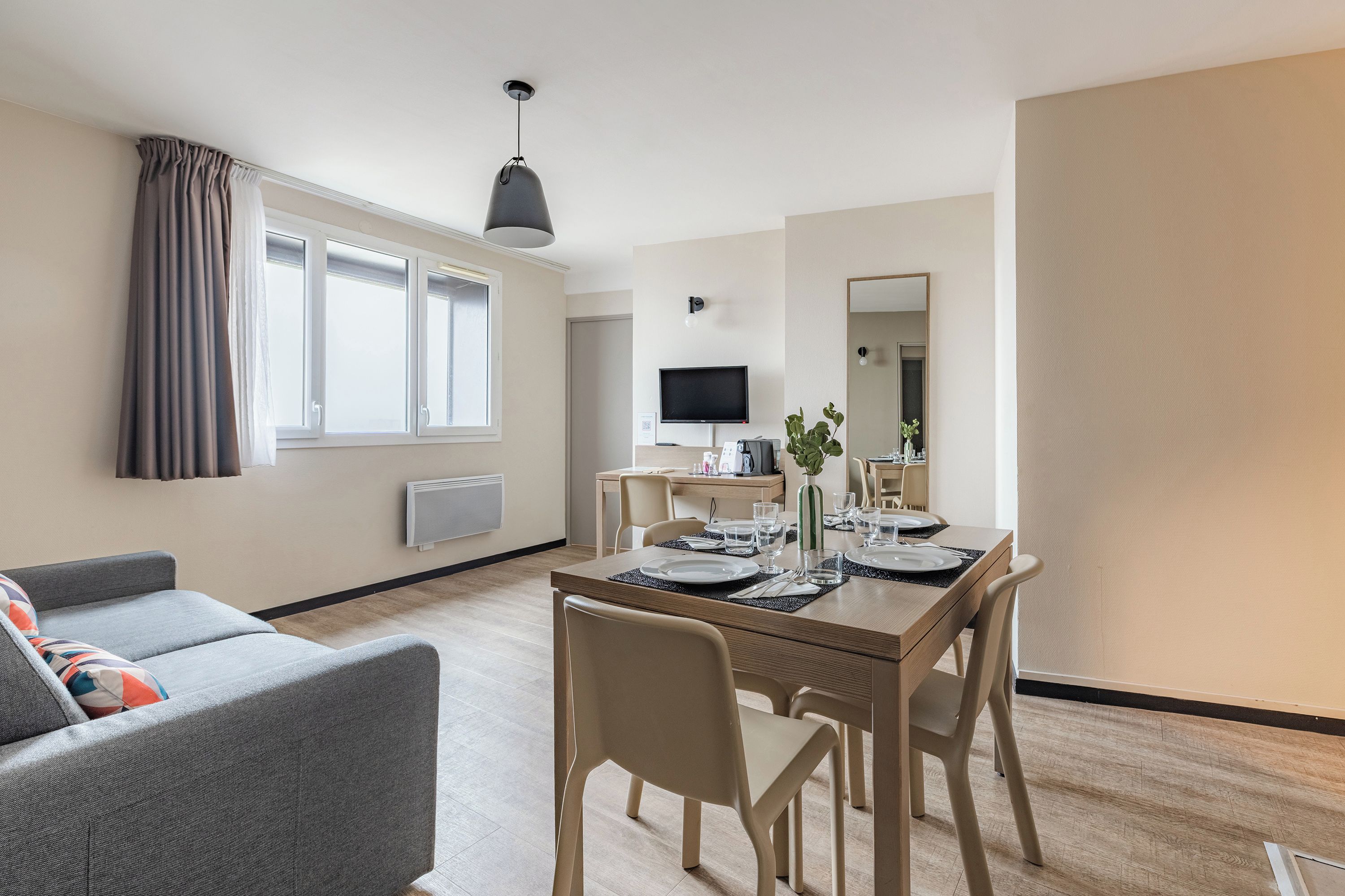 Un salon moderne avec un canapé gris, une table à manger dressée pour deux personnes et un téléviseur en arrière-plan.