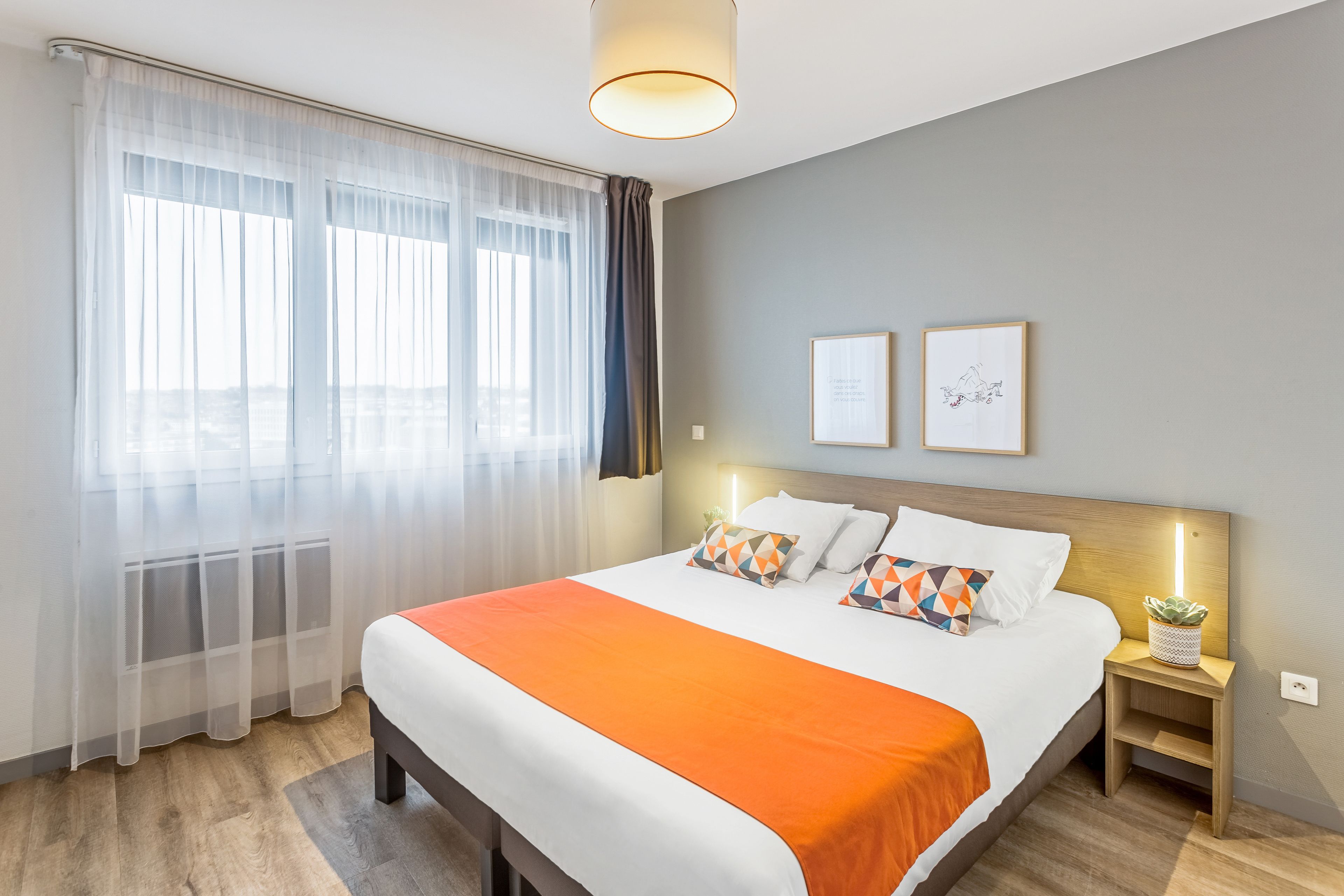 Ein Hotelzimmer mit einem großen Bett, einem Nachttisch mit Lampe und einem Fenster mit durchsichtigen Vorhängen.