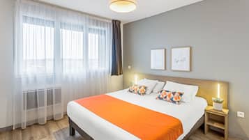 Ein Hotelzimmer mit einem großen Bett, einem Nachttisch mit Lampe und einem Fenster mit durchsichtigen Vorhängen.
