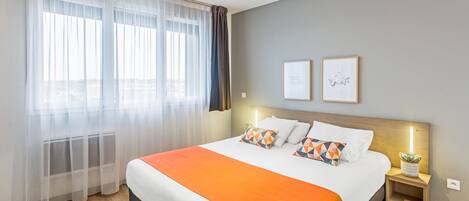 Una habitación de hotel con una cama grande, una mesita de noche con lámpara y una ventana con cortinas translúcidas.