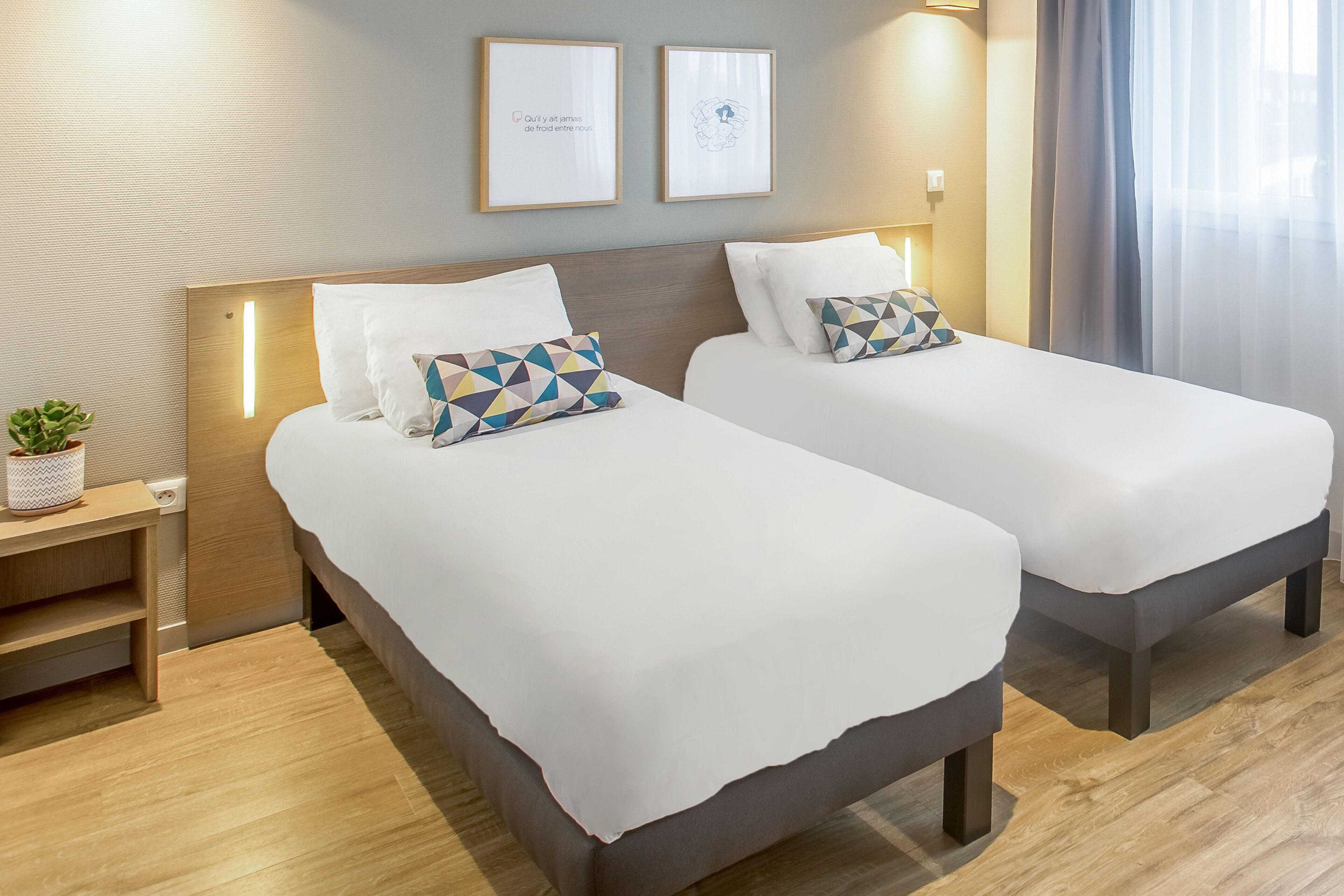 A hotel room with two beds, a bedside table with a plant, and framed artwork on the wall.