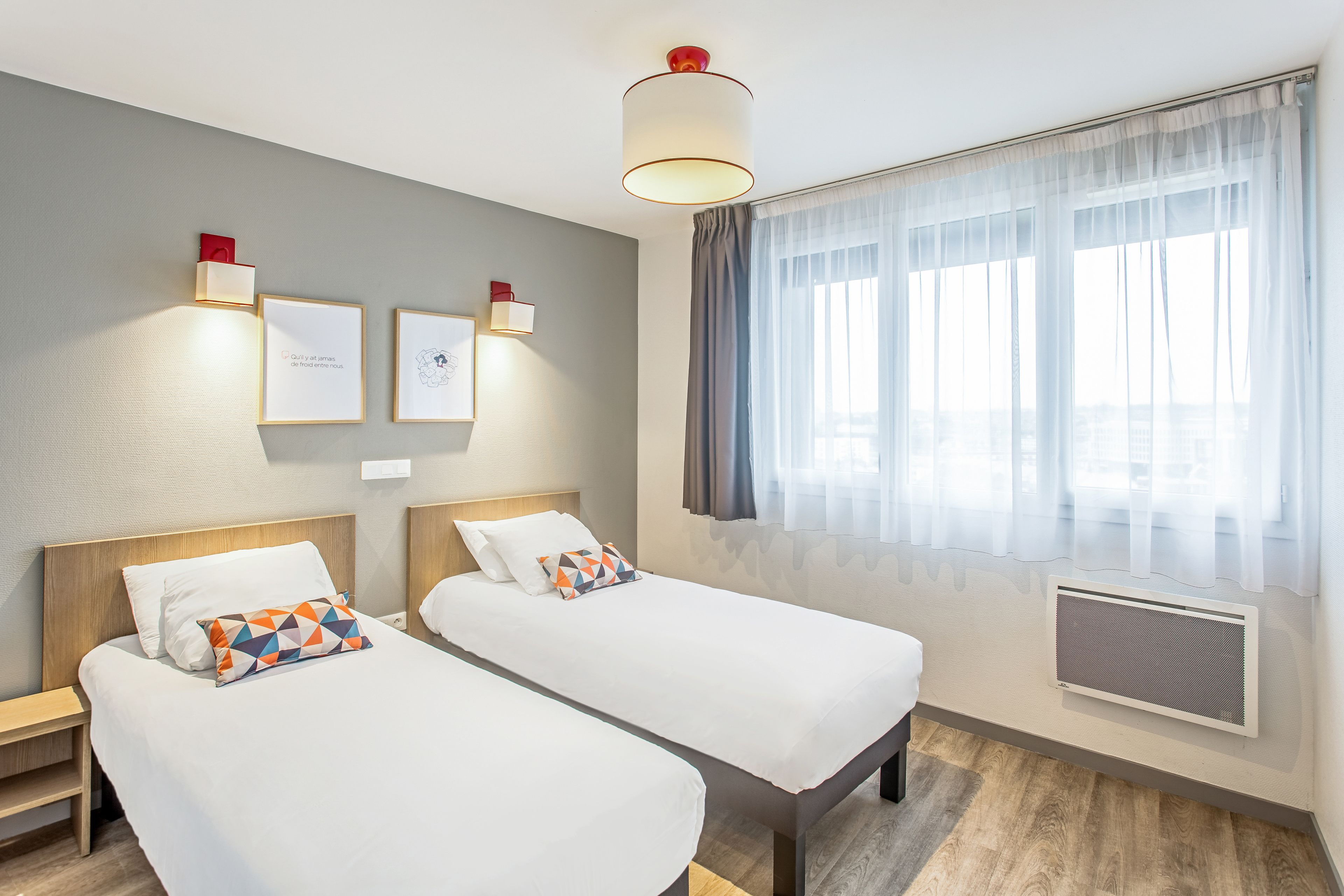 A hotel room with two beds, a window with sheer curtains, wall-mounted light fixtures, and framed artwork.