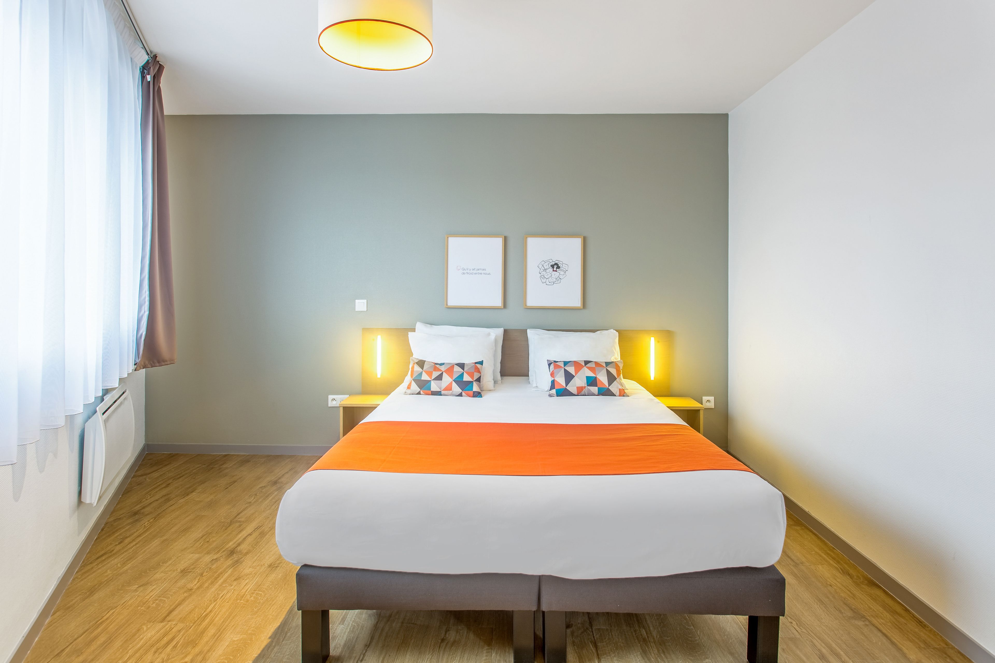 Una habitación de hotel moderna con una cama grande, dos mesitas de noche con lámparas, piso de madera y una ventana con cortinas.