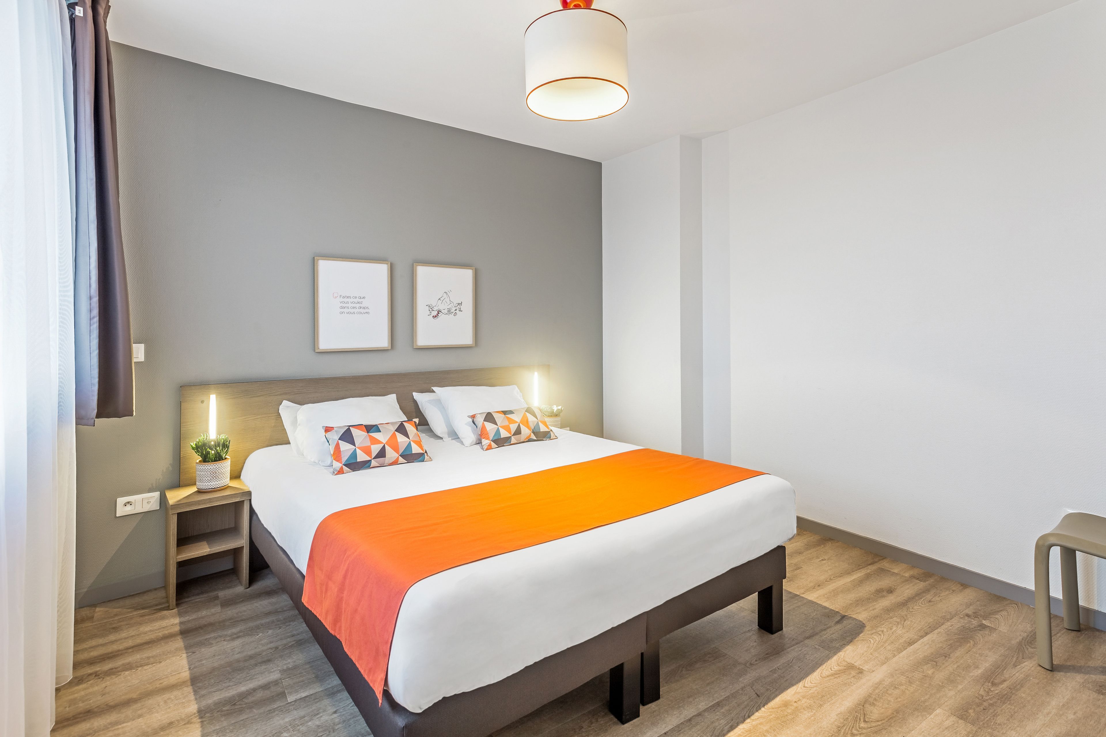 A modern hotel room with a large bed, a bedside table with a lamp, and a small plant.