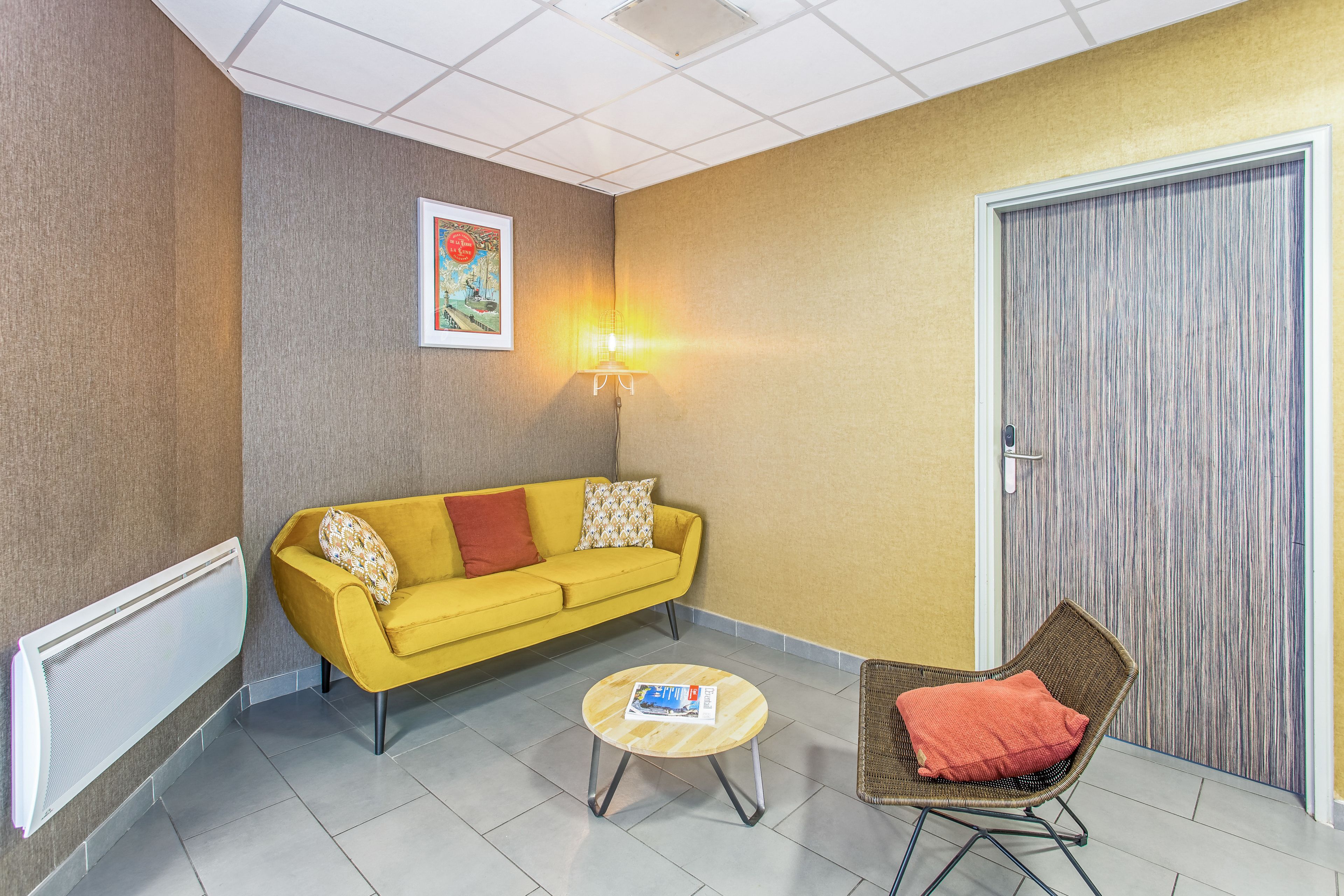 Een kamer met een gele bank, een rond houten bijzettafeltje, een rieten stoel, een wandlamp en een deur met een structuurpaneel.