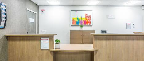 Een receptiegedeelte met houten balies, een kunstwerk van een stadsgezicht in een lijst, een potplant en een receptieteken.
