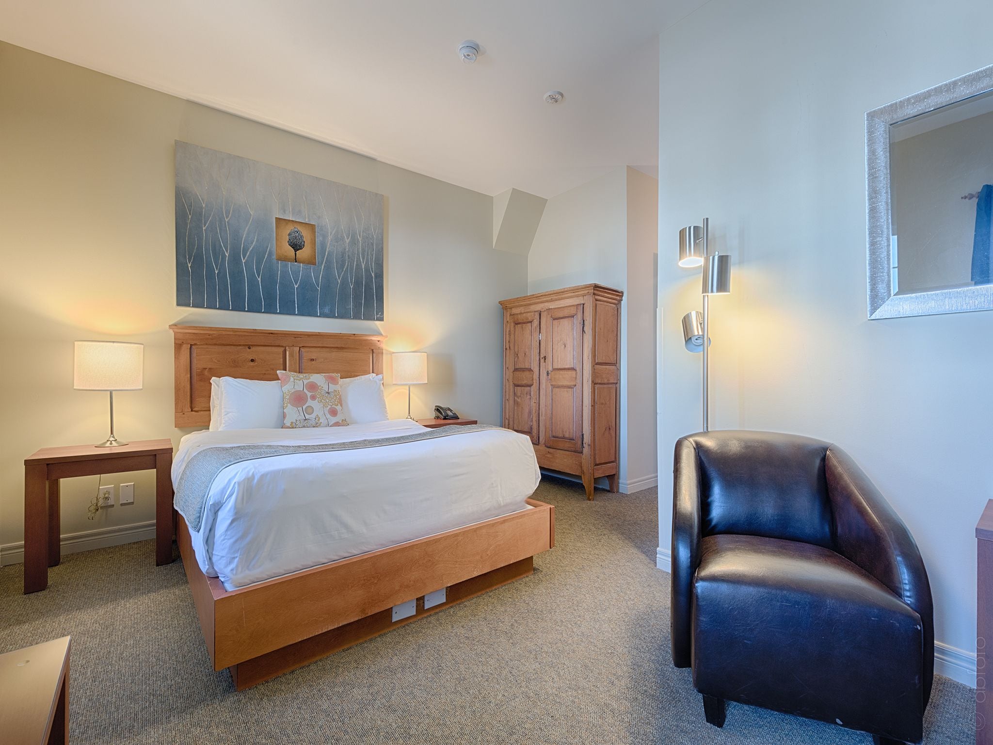 Una habitación de hotel con una cama grande, dos lámparas de noche, un armario de madera, un sillón de cuero y un cuadro en la pared.