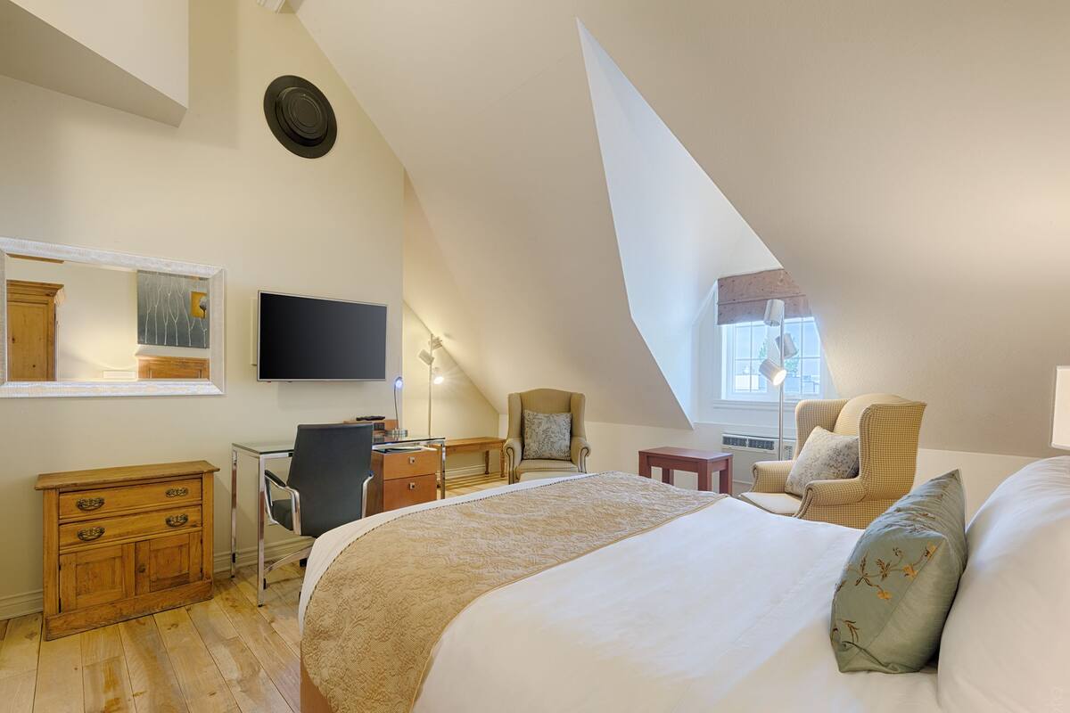 A bedroom with a large bed, a desk, a chair, a dresser, a television, and a window with natural light.