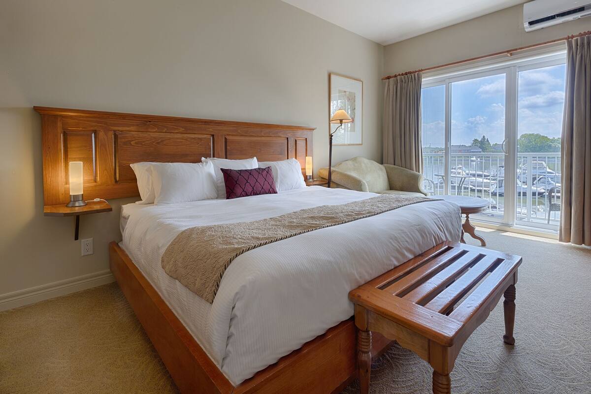 A hotel room with a large bed, a wooden headboard, a bedside table with a lamp, a chair, and a view of boats through large windows.