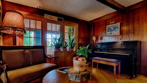 Una habitación acogedora con suelo de madera, un sofá, una mesa redonda con un libro y una cesta de manzanas, un piano y ventanas grandes con persianas.