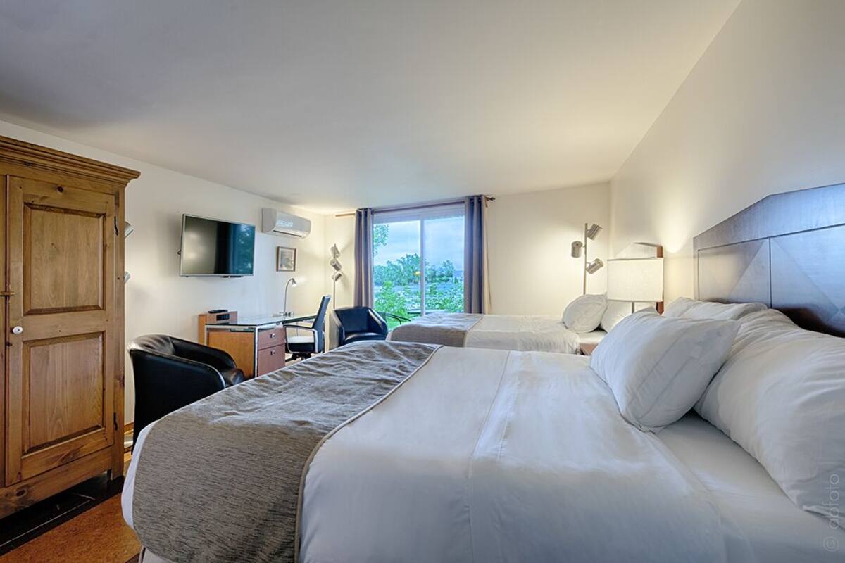 A hotel room with two beds, a desk, a chair, a TV, and a window with a view of greenery.