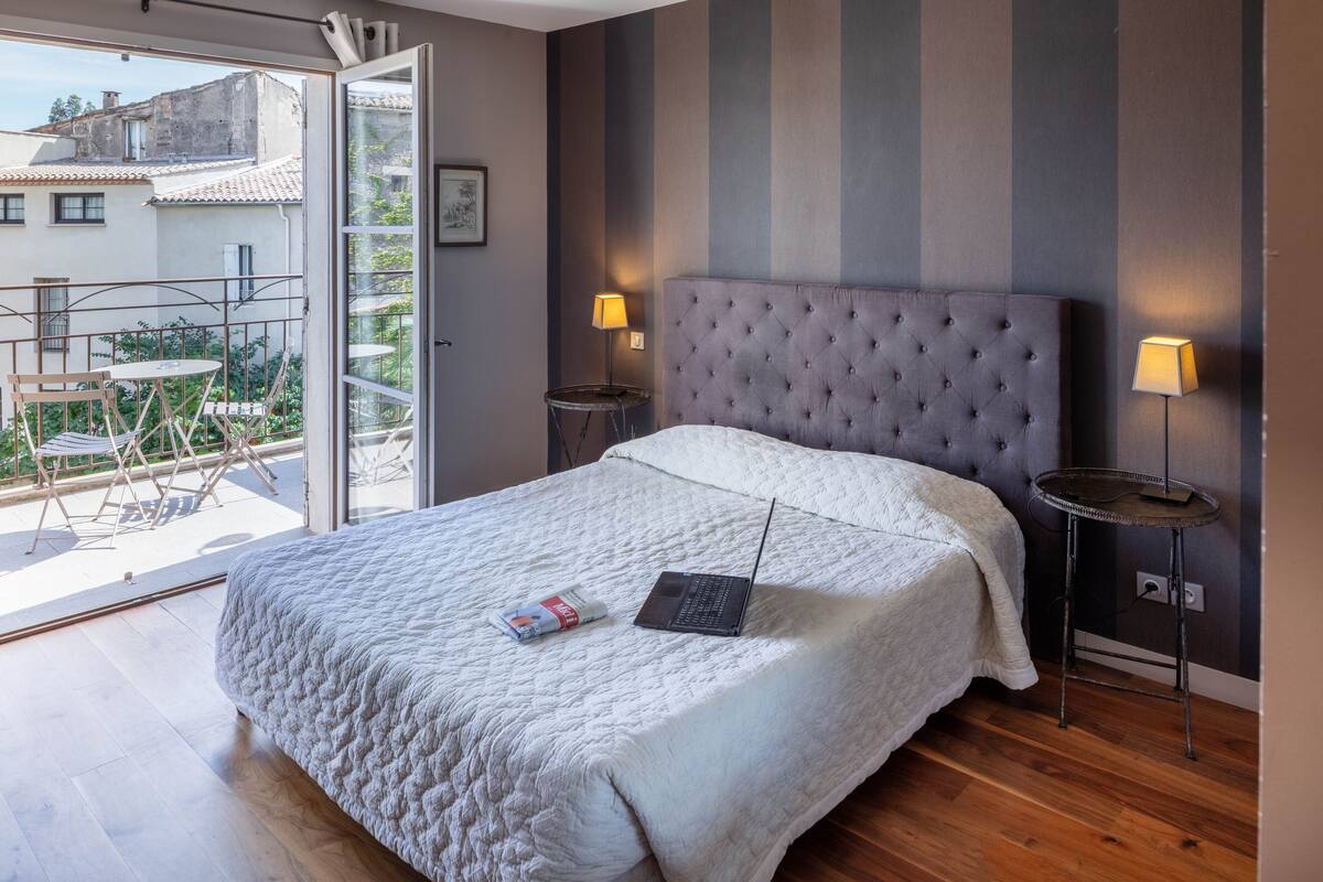 Un dormitorio con una cama grande, dos lámparas de noche, una computadora portátil sobre la cama y un balcón con mobiliario exterior.