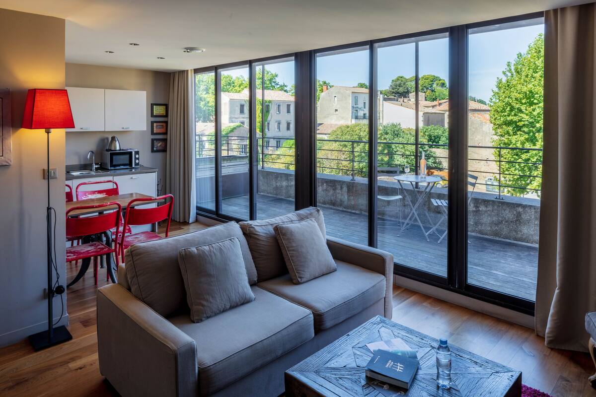 Una sala de estar moderna con un sofá, una mesa de centro y un comedor con sillas rojas. Hay una zona de cocina con un microondas y una lámpara roja. Amplios ventanales ofrecen vistas a edificios y árboles.