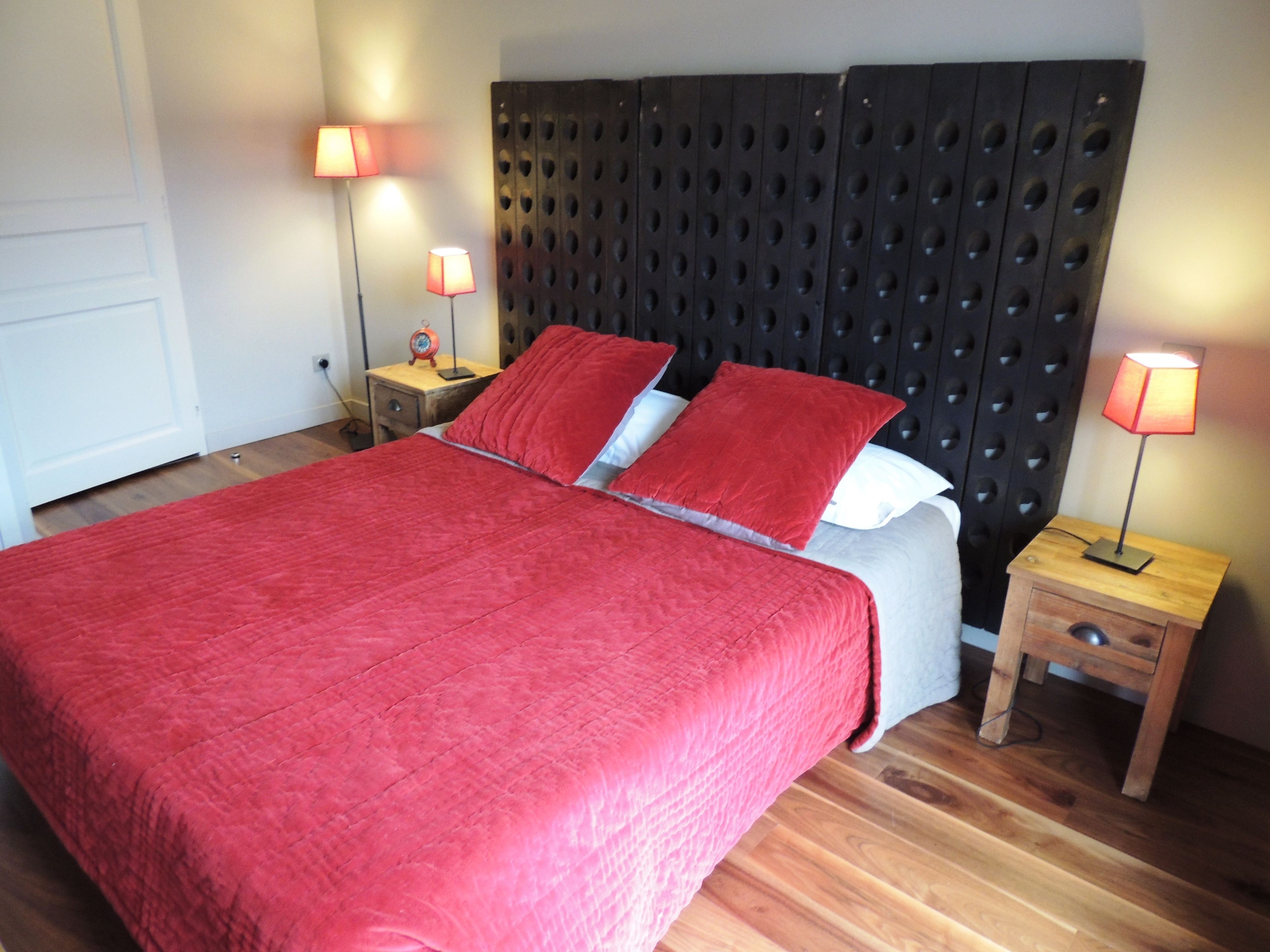 Una cama bien hecha con una colcha roja y almohadas, dos lámparas de noche, un mesita de noche de madera y un cabecero moderno.