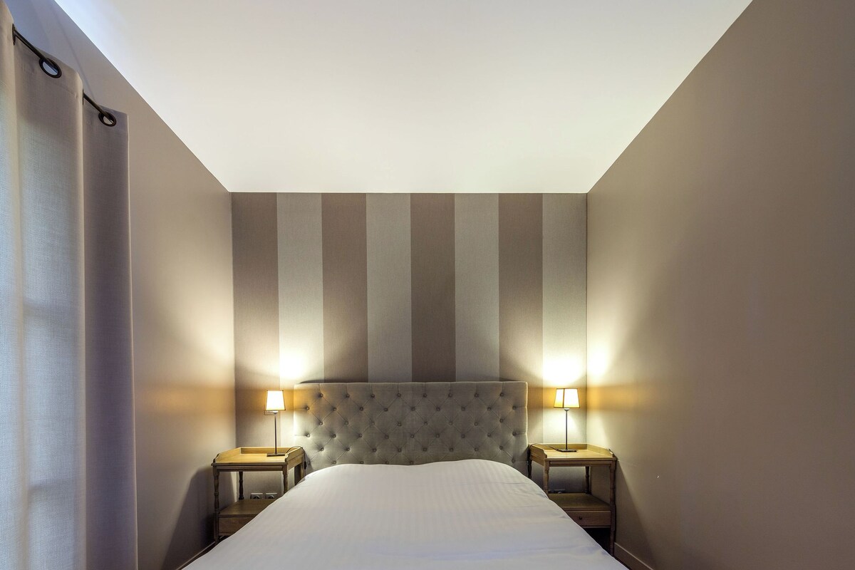 Un dormitorio con una cama grande, dos mesitas de noche con lámparas, suelo de madera y paredes a rayas.