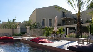 Un área de piscina con sillones de descanso y sombrillas, un edificio con balcones y una palmera.