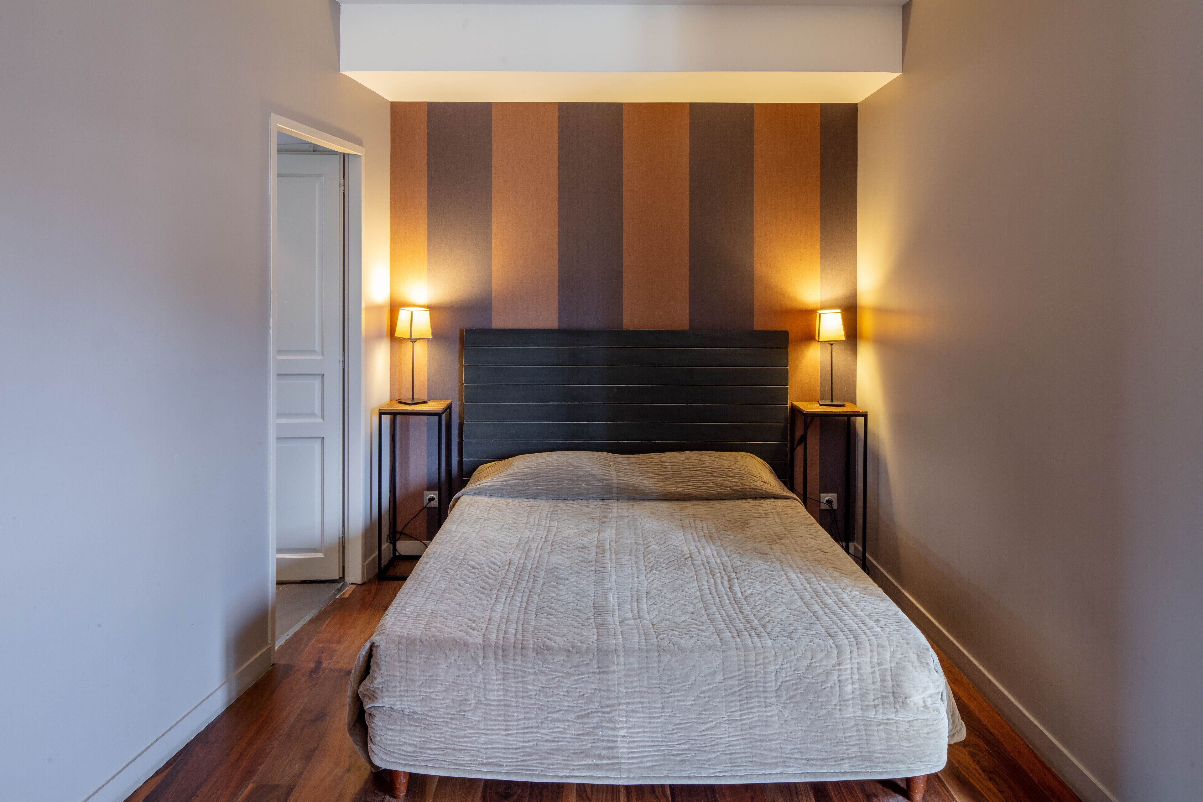 A neatly made bed with a textured blanket, flanked by two bedside lamps on wooden nightstands, in a room with a striped wall and a door on the left.