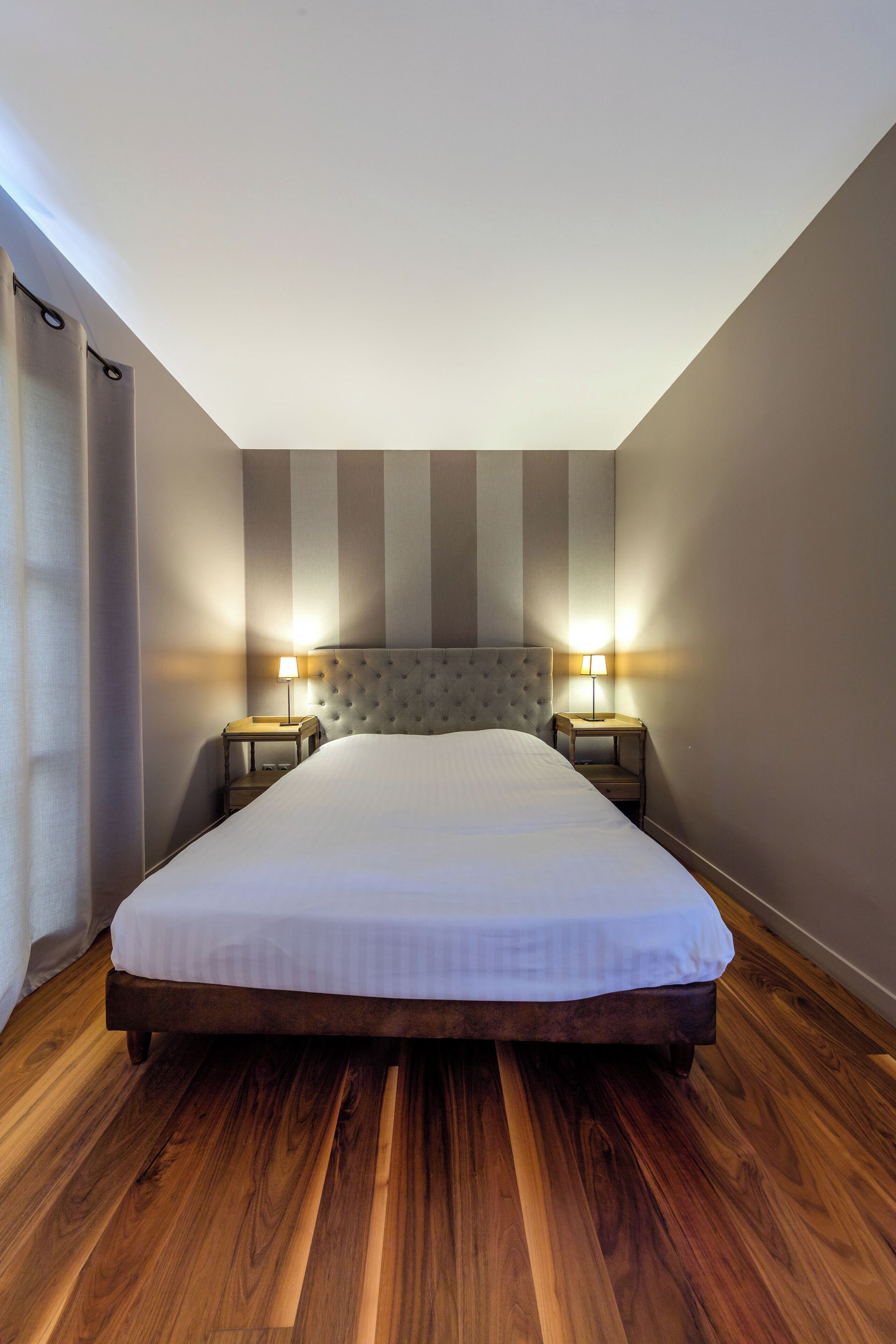 A bedroom with a large bed, two bedside tables with lamps, wooden flooring, and striped walls.