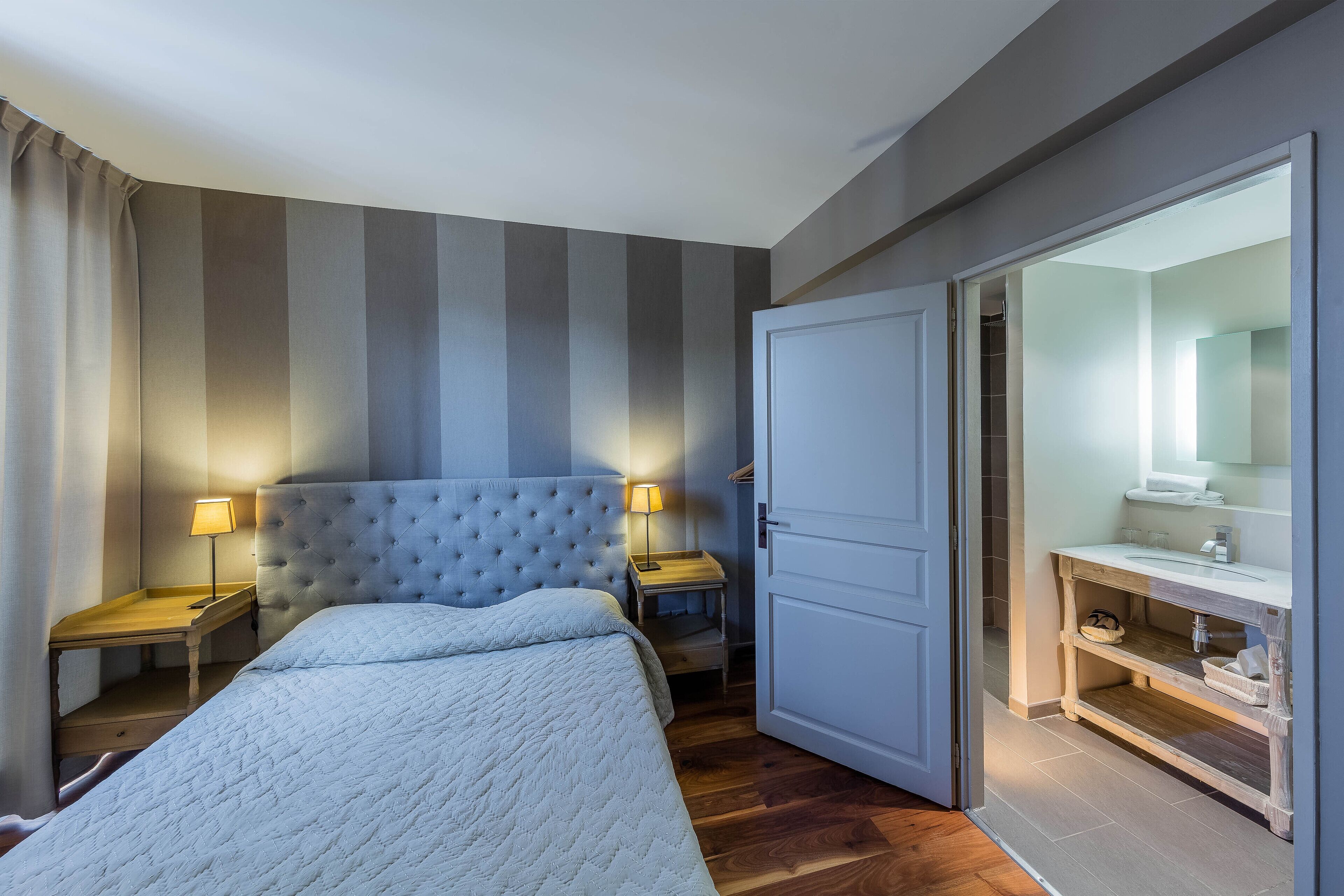 A hotel room with a bed, nightstands, a lamp, a wardrobe, and a bathroom with a mirror and sink.