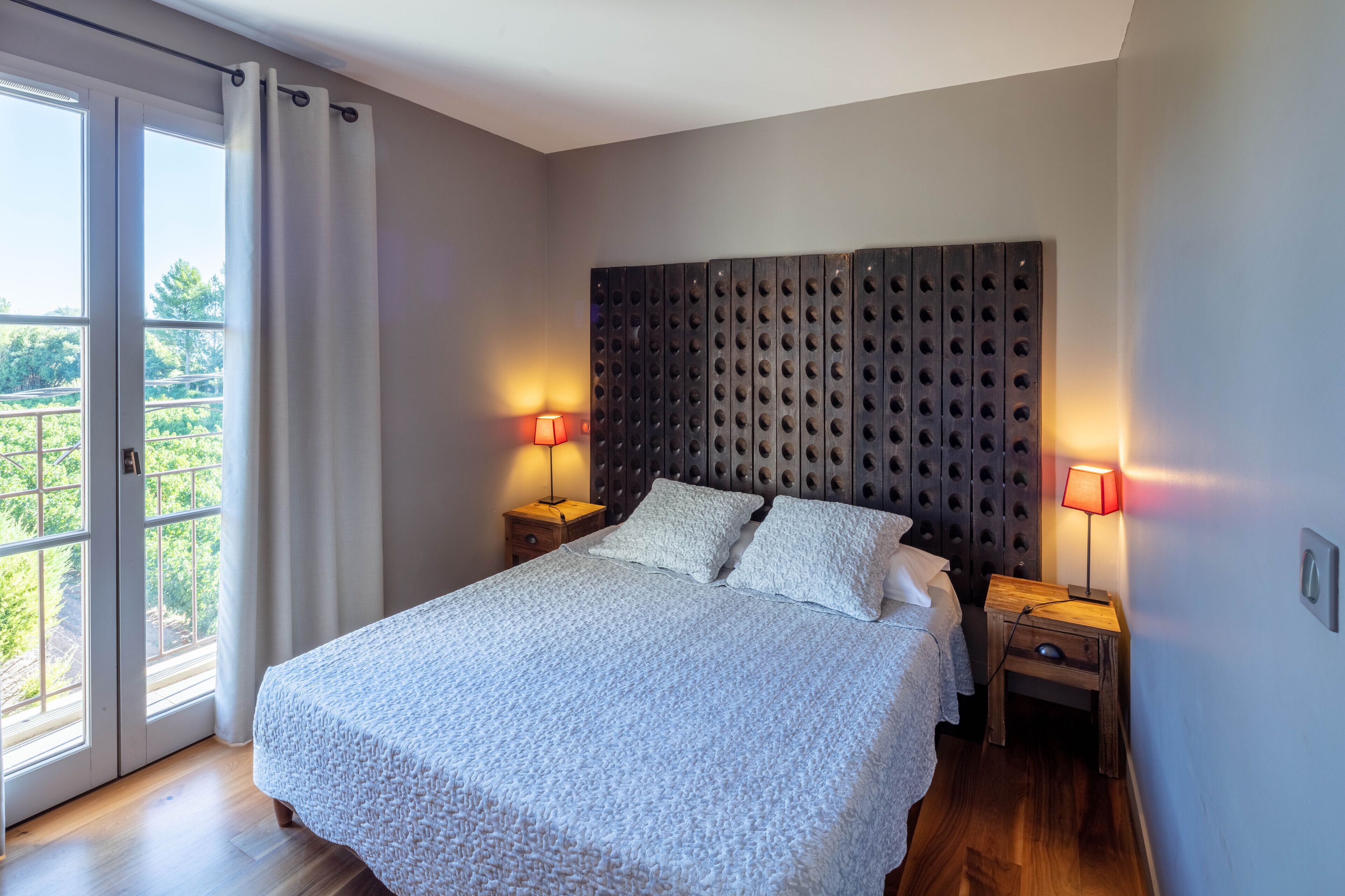 A bedroom with a large bed, two bedside tables with lamps, a dark headboard, and a view through a sliding glass door.