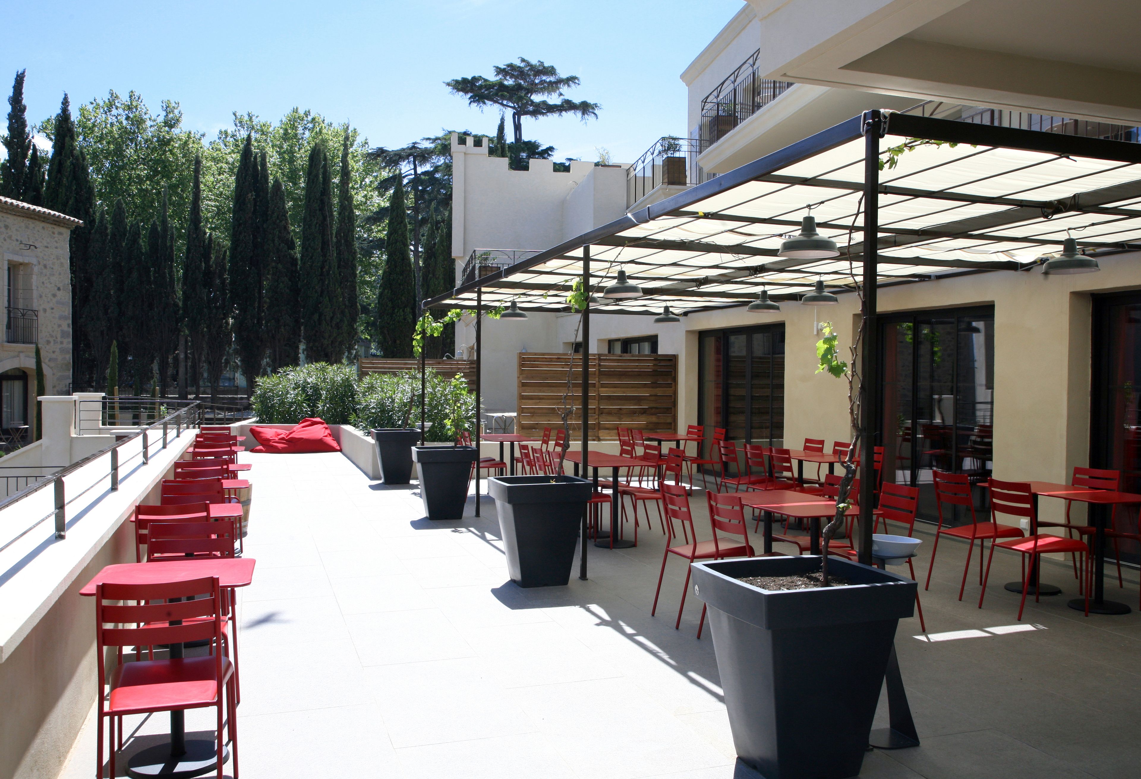 An outdoor patio with red tables and chairs, large planters, and a covered seating area with a pergola.