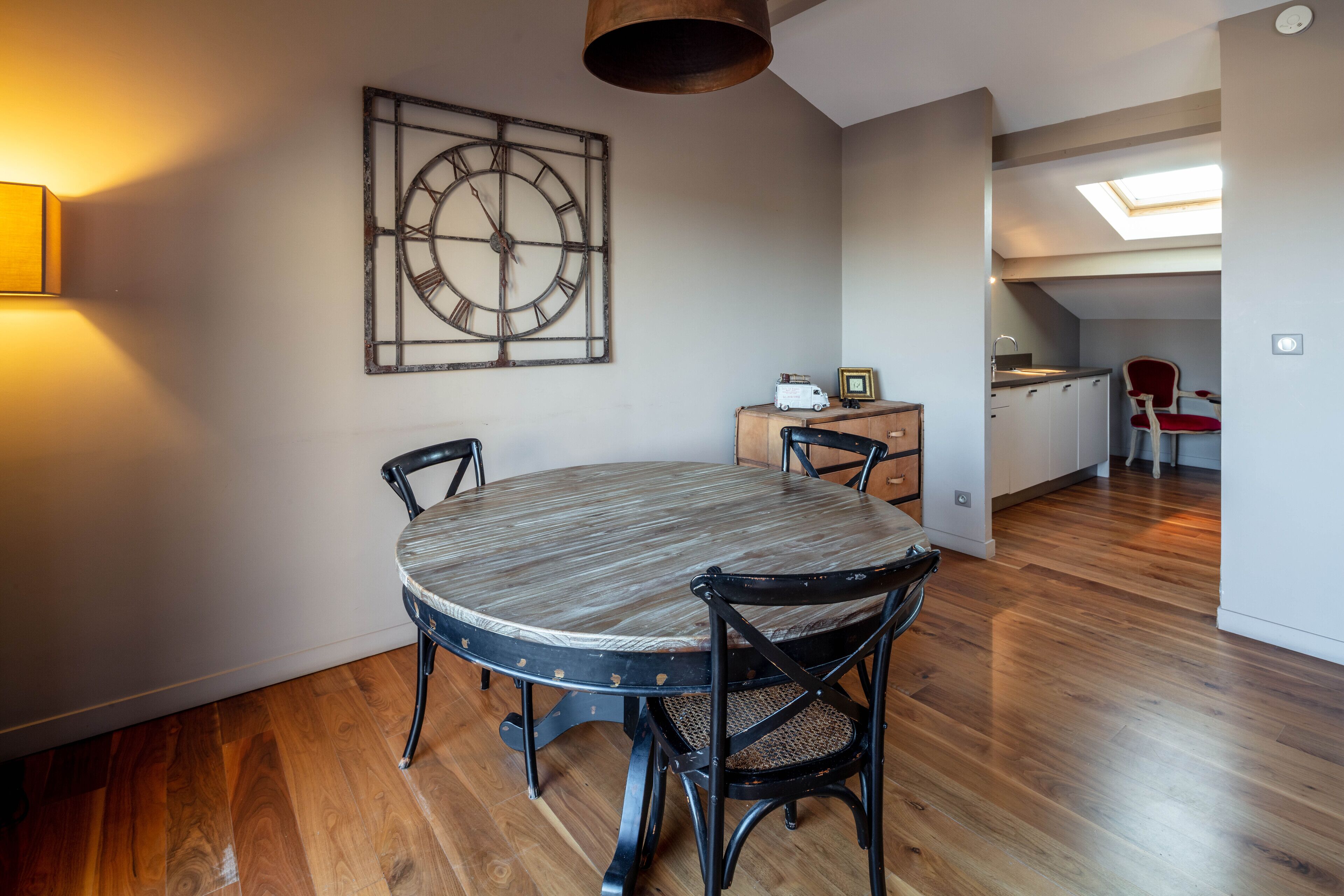 Un coin repas avec une table ronde et quatre chaises, une horloge murale, un petit meuble contenant un jouet et une lucarne au-dessus d’un espace cuisine.