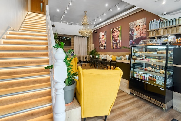 A staircase with wooden steps and white balusters, a yellow armchair, a chandelier, a wall with cultural artwork, and a display case with baked goods.