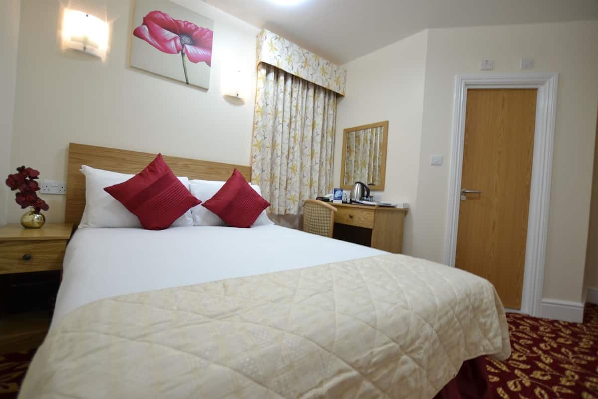 A hotel room with a bed, a desk, a chair, a nightstand, a vase with flowers, a painting, a mirror, and a door.