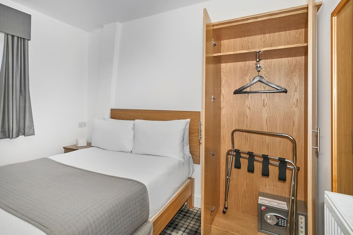 A hotel room with a bed, a wooden wardrobe, a coat rack, and a thermostat.