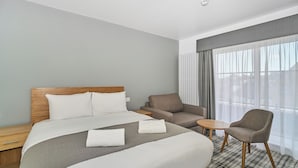 A hotel room with a large bed, two armchairs, a small table, and a view of the city through sheer curtains.