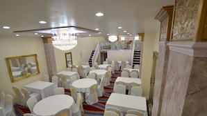 A banquet hall with round tables, white chairs, chandeliers, and a staircase.