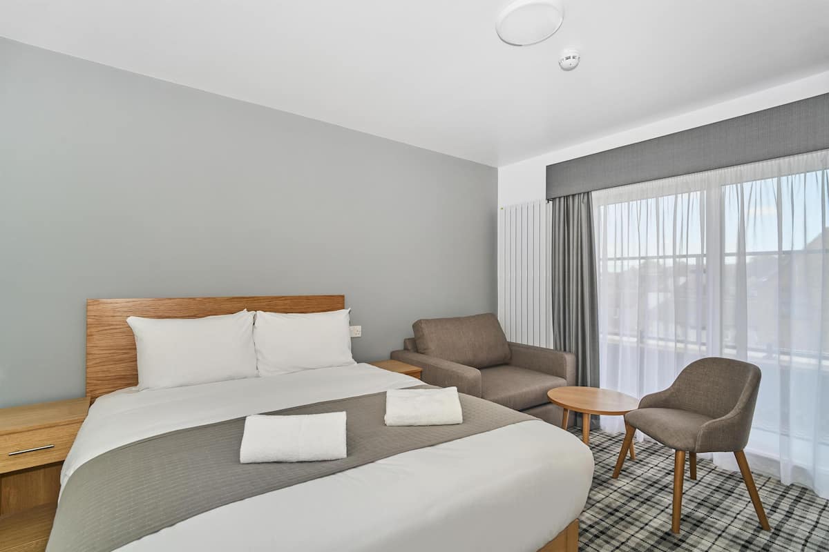 A hotel room with a large bed, two armchairs, a small round table, and a view of the city through sheer curtains.
