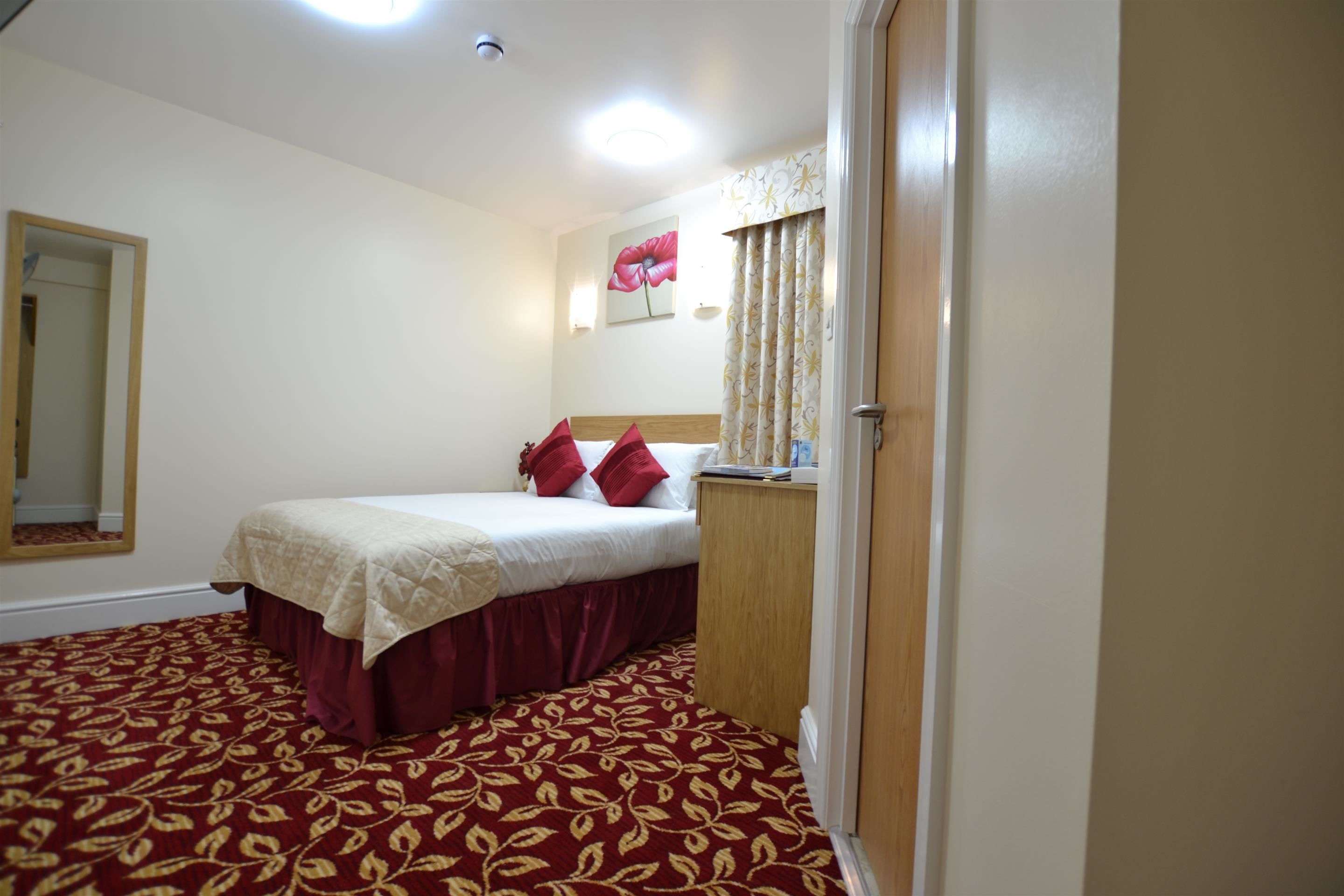 A hotel room with a bed, a wooden nightstand, a mirror, a red patterned carpet, and a wall with a floral painting.