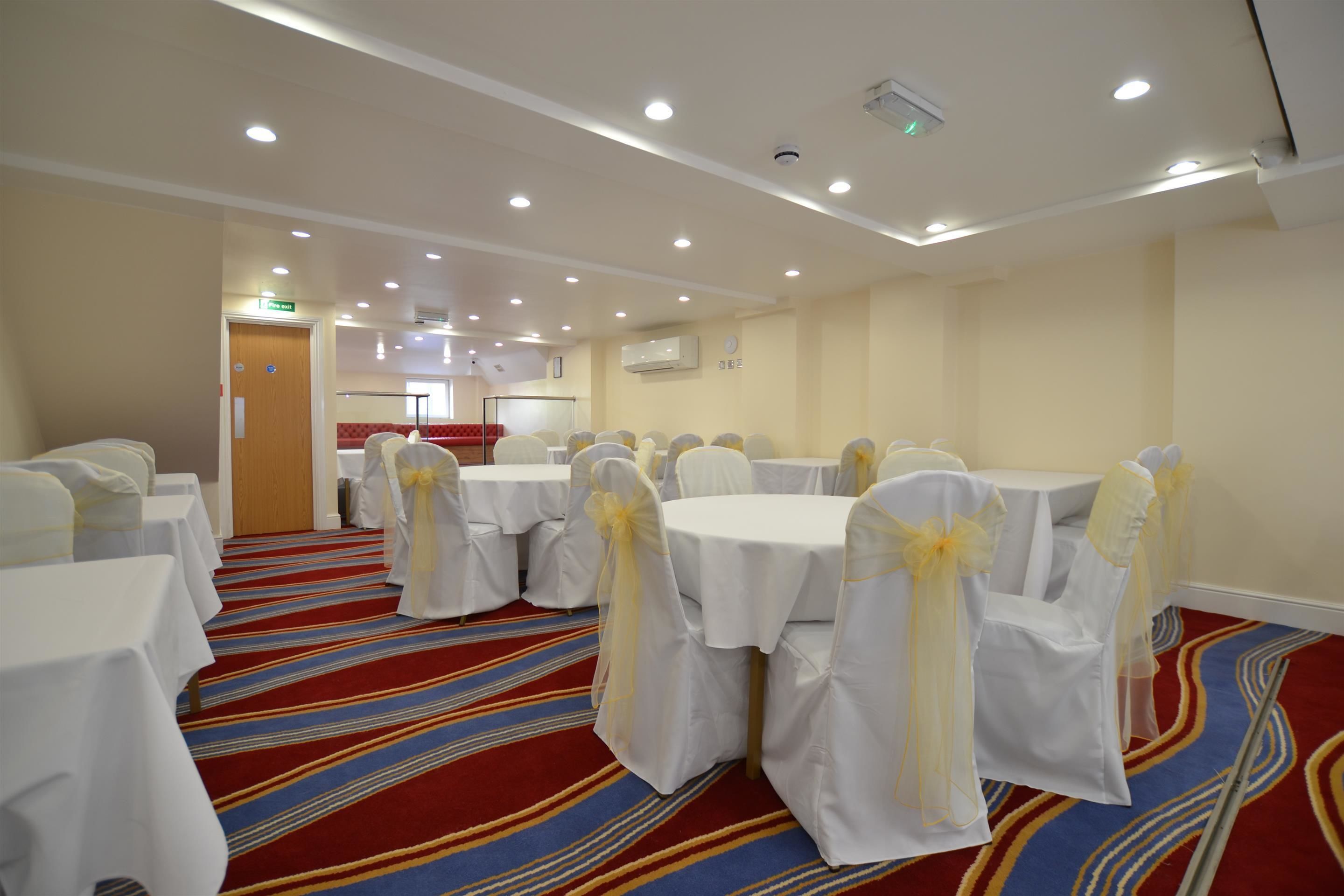A conference room with round tables, chairs with white covers and yellow sashes, a red and blue striped carpet, and recessed ceiling lighting.