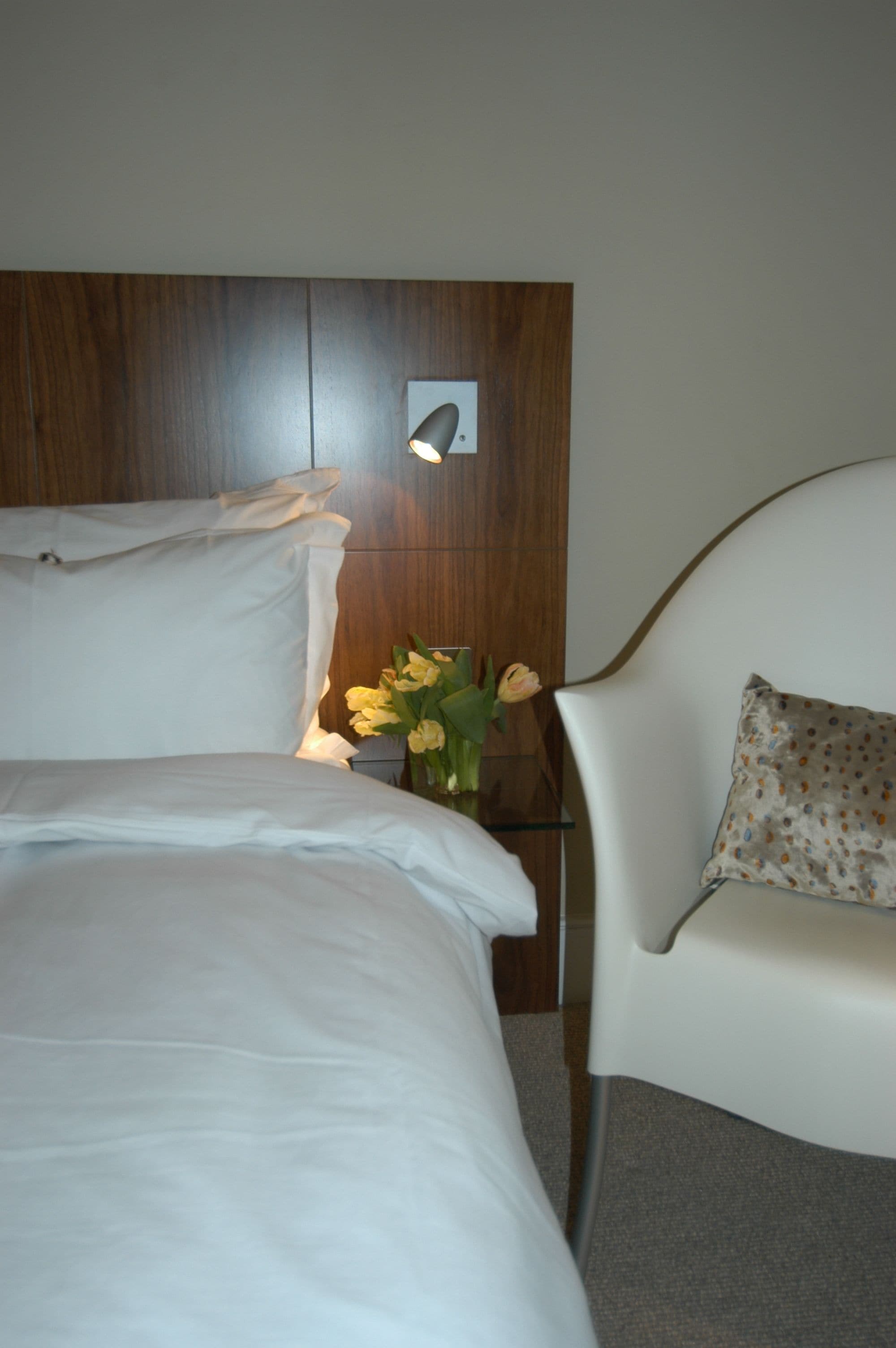 Una cama bien hecha con sábanas blancas, una mesita de noche con lámpara y una silla con cojín estampado.
