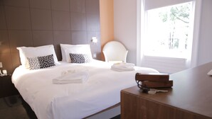 Una cama bien tendida con sábanas blancas y dos almohadas, una mesita de noche de madera con un teléfono y un libro, y una ventana con persianas.