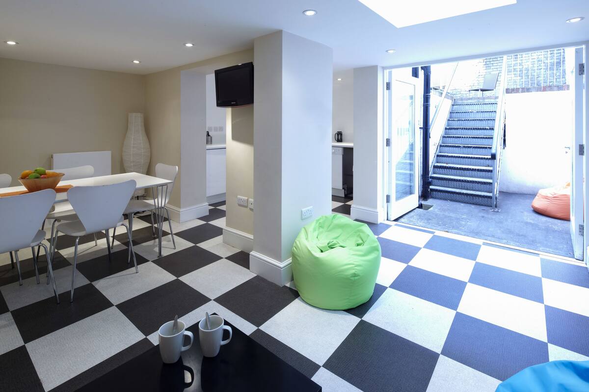 A modern living room with a checkered floor, a green bean bag, a dining area with a white table and chairs, and a TV mounted on the wall.