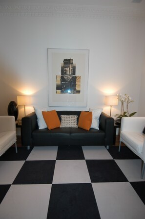 A living room with a black and white checkered floor, a black sofa with orange and white cushions, two white armchairs, and a framed abstract painting on the wall. - B+B Belgravia (London)