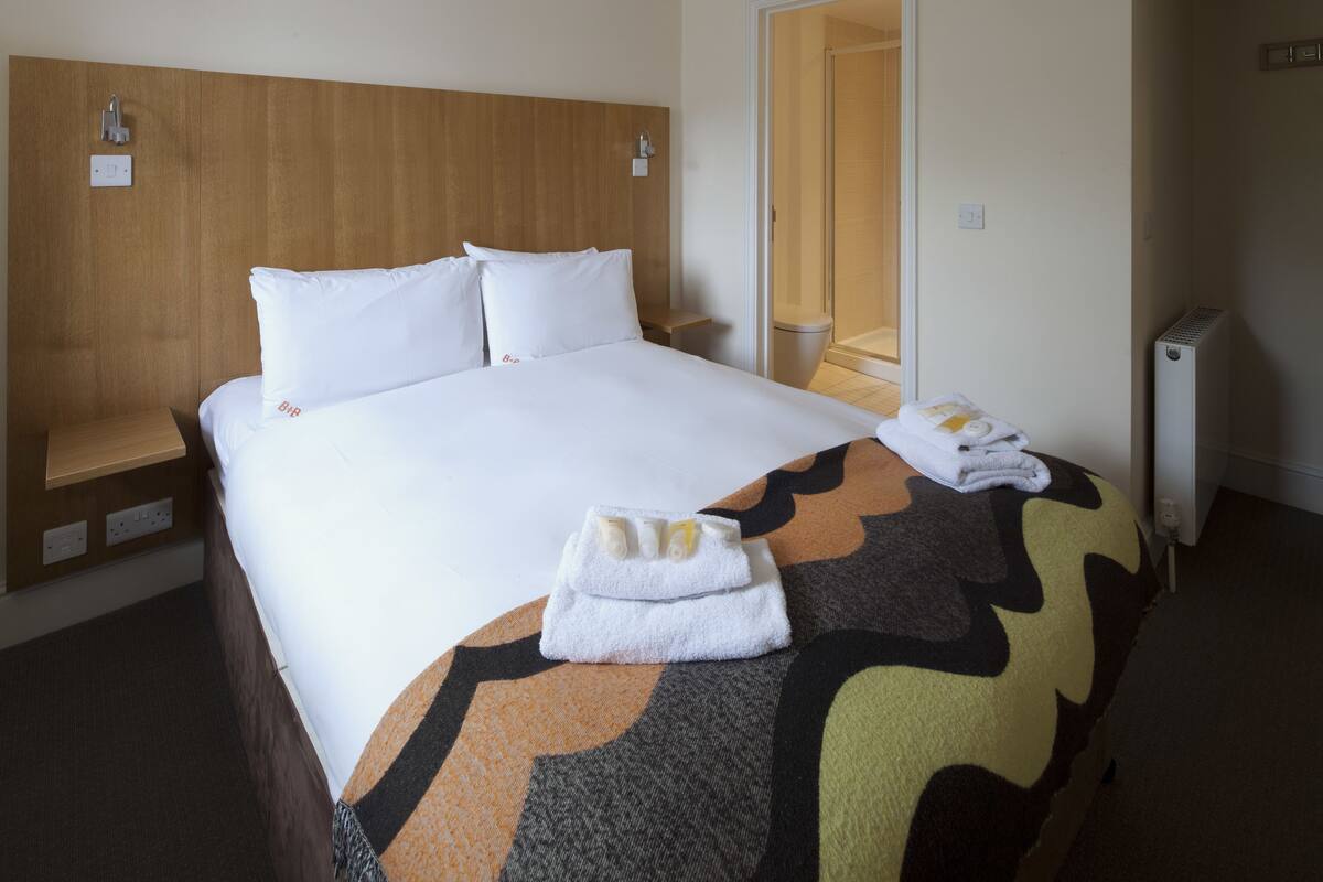 A hotel room with a large bed, a wooden headboard, a bedside table, a shower area, and a radiator.