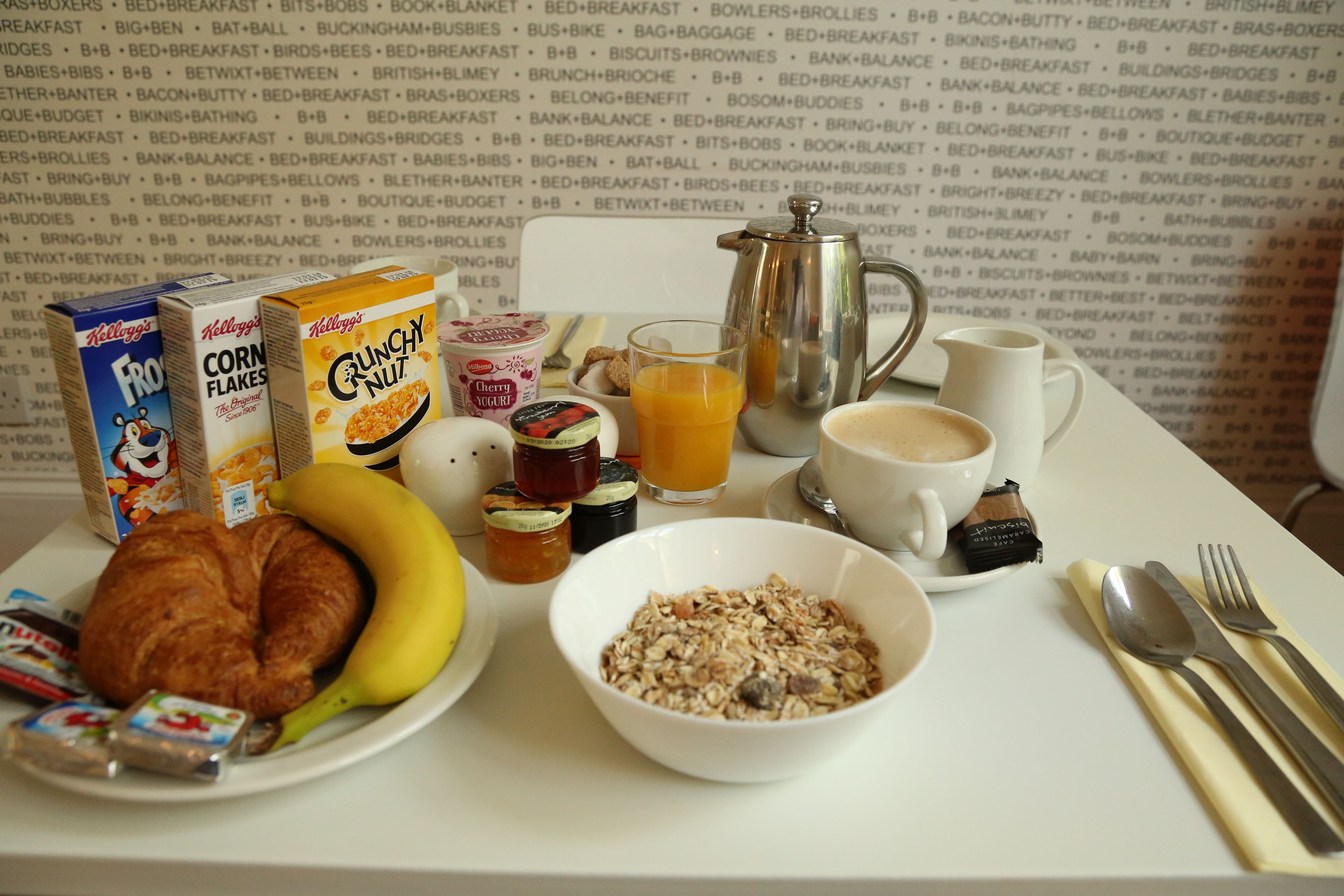 A breakfast table with cereal, a banana, a croissant, a bowl of granola, a coffee cup, and a teapot.
