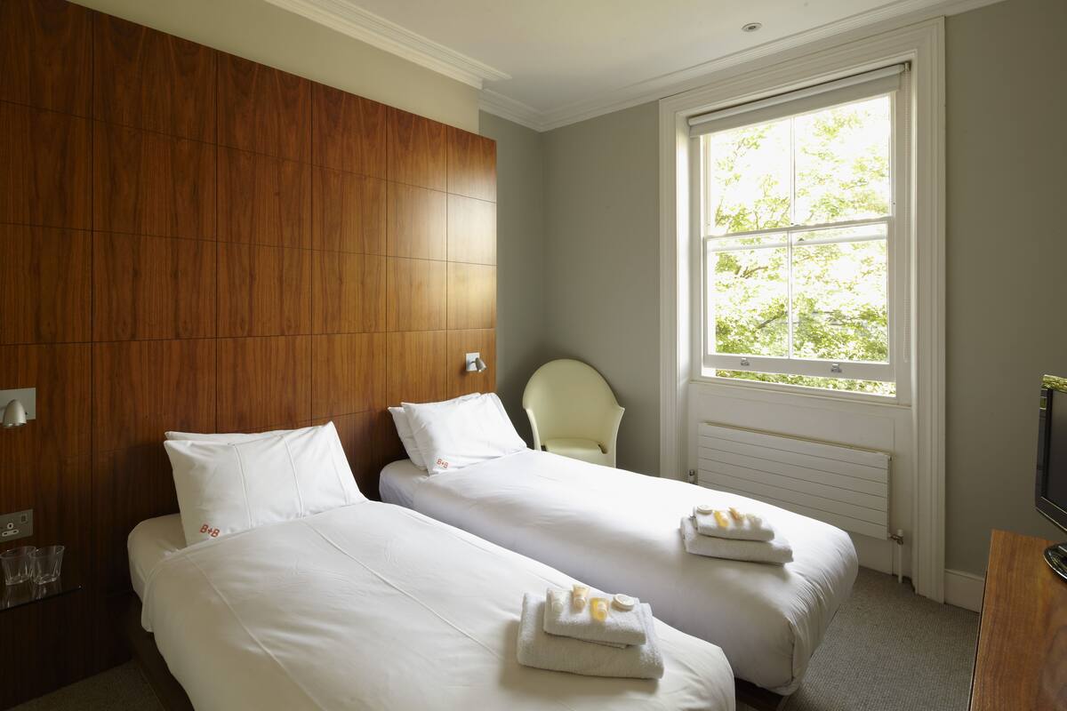 A hotel room with two beds, a wooden headboard, a window with a view of trees, and a chair.