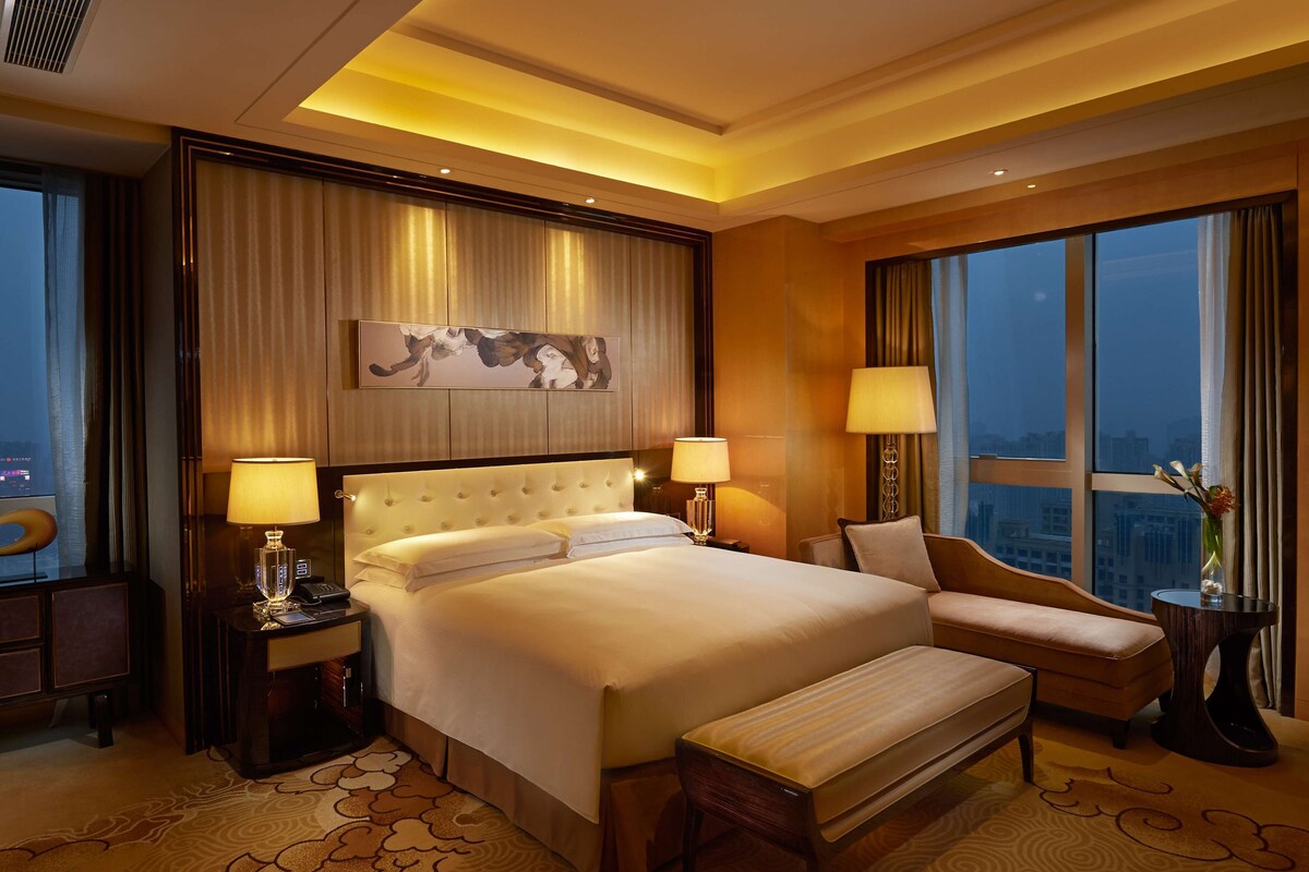A hotel room with a large bed, two bedside tables with lamps, a bench, a chair, a large window with a city view, and a decorative wall piece.