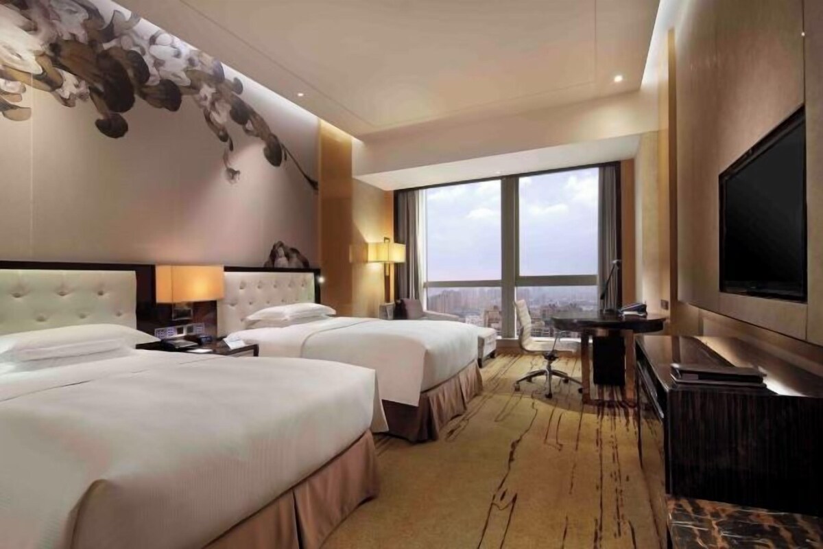 A hotel room with two beds, a desk, a TV, and a large window offering a city view.