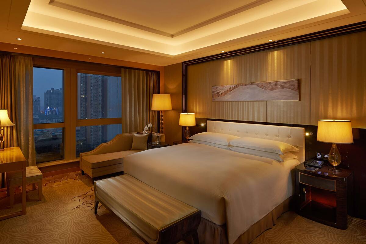 Une chambre d’hôtel dotée d’un grand lit, de deux tables de chevet avec des lampes, d’un banc et offrant une vue sur le paysage urbain grâce à de grandes fenêtres, ainsi qu’un design moderne au niveau du plafond.